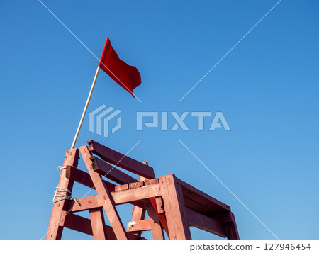 Red flag on a wooden platform. Flag as a symbol of danger. Bathing is prohibited. Beach on the sea. The concept of rescue work. Red flag on a wooden platform. Flag as a symbol of danger. Bathing is prohibited. Beach on the sea. The concept of rescue work. 127946454