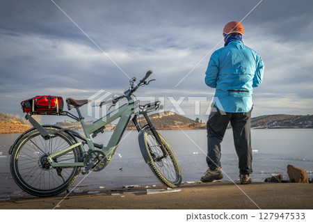 Male cyclist with a touring e-bike on a shore of frozen Horsetooth Reservoir in Colorado foothills. Male cyclist with a touring e-bike on a shore of frozen Horsetooth Reservoir in Colorado foothills. 127947533