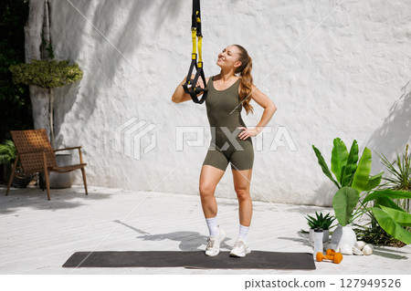 Smiling woman training with suspension straps outdoors promoting fitness healthy lifestyle and functional bodyweight exercise 127949526