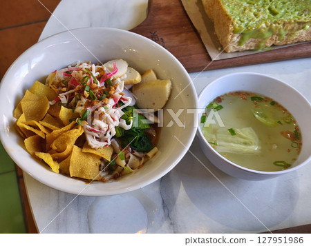 a vibrant bowl of Asian noodles, topped with crispy wonton skins, fish cakes, green vegetables, and colorful garnishes. a vibrant bowl of Asian noodles, topped with crispy wonton skins, fish cakes, green vegetables, and colorful garnishes. 127951986
