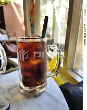Lo Han Kuo or Monk Fruit Drink, an iced herbal beverage with a dark amber hue and a spoon, set by a sunlit window. 127952128