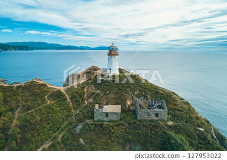 Lighthouse stands on rocky coastline, overlooking the ocean with mountains in the background during a clear day Lighthouse stands on rocky coastline, overlooking the ocean with mountains in the background during a clear day 127953022