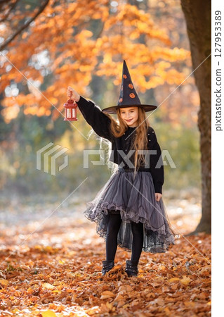 Cute happy little girl dressed in witch costume standing with over autumn forest background. Girl wearing dress and cone hat and holding a lantern. Girl dressed for Halloween. 127953389