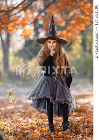 Cute happy little girl dressed in witch costume standing with over autumn forest background. Girl wearing dress and cone hat. Girl dressed for Halloween. 127953390