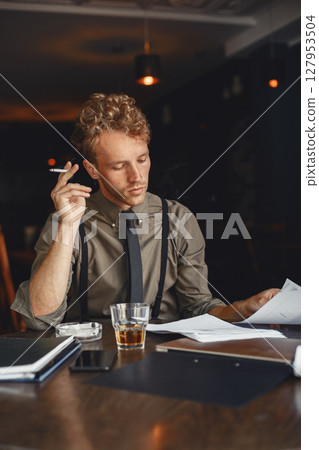 Man drinks whiskey. Businessman reads documents. Director in a shirt and suspenders. 127953504