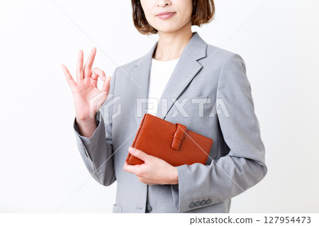 A middle-aged woman in a suit holding a notebook and making a circle sign A middle-aged woman in a suit holding a notebook and making a circle sign 127954473