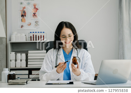 Confident young Asian female doctor in white medical uniform sit at desk working on computer. Smiling use laptop write in medical journal Confident young Asian female doctor in white medical uniform sit at desk working on computer. Smiling use laptop write in medical journal 127955838