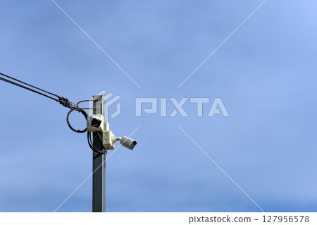 A high-angle surveillance camera mounted on a steel pole contrasts against the backdrop of a clear sky, evoking both vigilance and stillness. 127956578