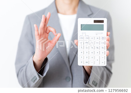 A middle-aged woman in a suit holding a white calculator and making a circle sign A middle-aged woman in a suit holding a white calculator and making a circle sign 127956692