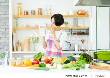 A middle-aged housewife wearing an apron holding a calculator in the kitchen A middle-aged housewife wearing an apron holding a calculator in the kitchen 127956882