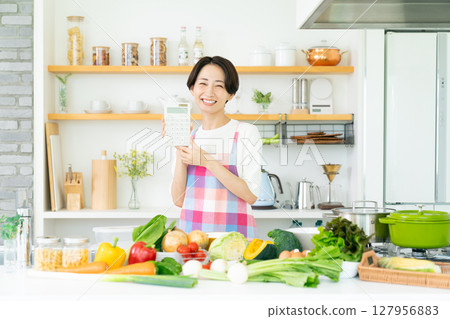 A middle-aged housewife wearing an apron holding a calculator in the kitchen A middle-aged housewife wearing an apron holding a calculator in the kitchen 127956883