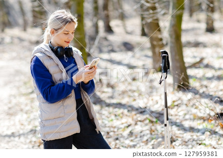 Young hiker woman using smartphone and listening music with headphones and trekking poles in forest Young hiker woman using smartphone and listening music with headphones and trekking poles in forest 127959381