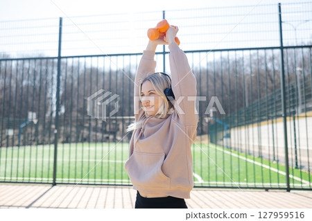 Young sportswoman lifting dumbbells and listening to music with headphones during outdoor workout 127959516
