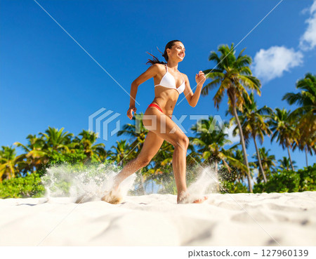 AI generated dynamic low angle shot of woman in red and white bikini running across the camera with sand kicking up, palm trees in background under bright blue sky creating energetic summer atmosphere 127960139