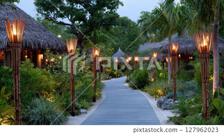 A walkway lined with tiki torches illuminates a tropical resort, creating a serene evening atmosphere. Vacationers stroll along the path to enjoy the tropical getaway 127962023