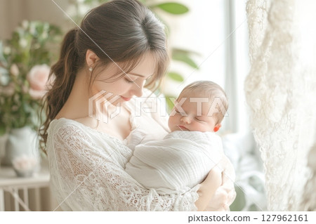 Young mother in white lace dress tenderly holds sleeping newborn baby near window with soft natural light 127962161