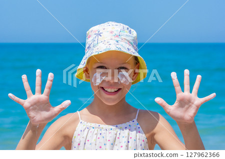 Happy girl at the beach, wearing a colorful sun hat, with sunscreen applied on her face and hands raised, enjoying summer fun and promoting sun safety Happy girl at the beach, wearing a colorful sun hat, with sunscreen applied on her face and hands raised, enjoying summer fun and promoting sun safety 127962366