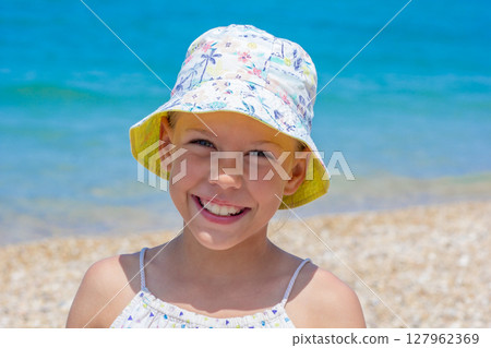 Smiling girl in a vibrant sun hat enjoys a sunny day at the beach, with soft sand and clear turquoise water in the background 127962369