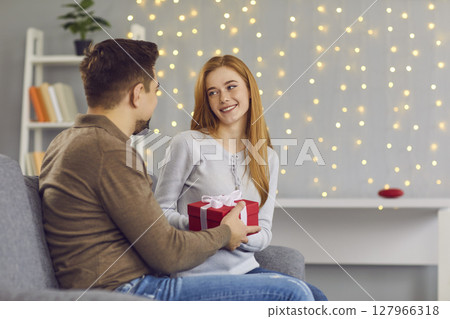 Young smiling woman girlfiend sitting and getting present box with ribbon from loving man Young smiling woman girlfiend sitting and getting present box with ribbon from loving man 127966318