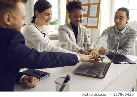 Multiracial diverse business team putting hand together sitting at office desk Multiracial diverse business team putting hand together sitting at office desk 127966420