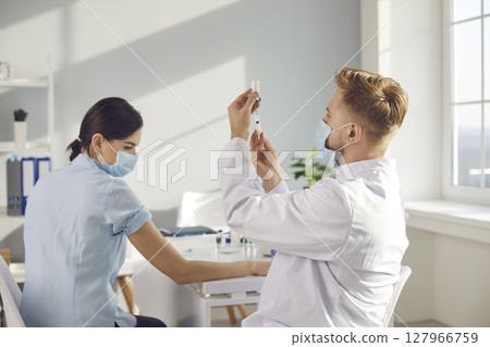 Young man doctor in mask and uniform preparing syringe for making vaccination Young man doctor in mask and uniform preparing syringe for making vaccination 127966759