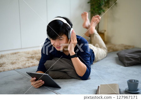 Happy man lying on the floor with headphones watching content on a digital tablet in a living room 127967171