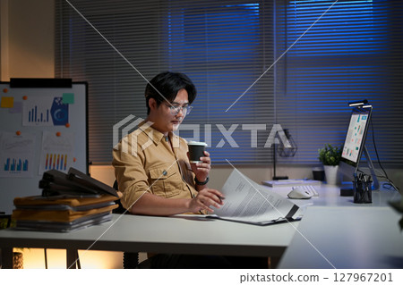 Focused man in glasses holding coffee while working overtime in a modern office Focused man in glasses holding coffee while working overtime in a modern office 127967201