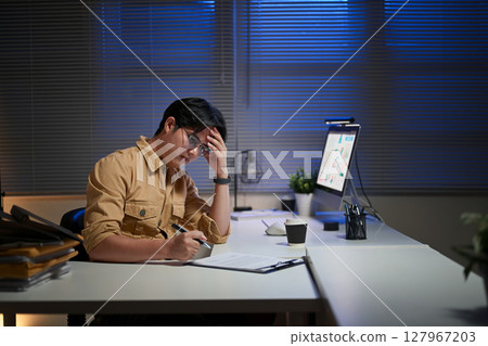 Tired employee holding his head in frustration while reviewing documents late at night in an office 127967203