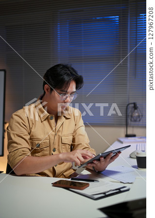 Focused office worker analyzing data on digital tablet during overtime hours in an office 127967208