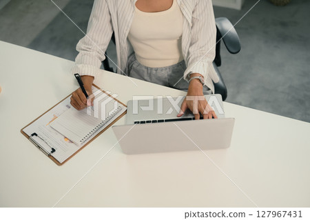 Top view of female entrepreneur using laptop and taking notes on clipboard 127967431