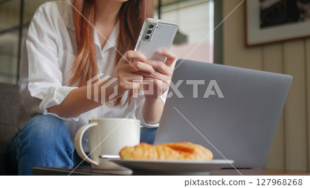 front view of woman using phone and laptop computer at coffee shop for work in the morning with hot coffee, croissant bakery and mobile phone on the table. anonymous face 127968268