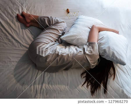 woman lying on bed, cover her head with pillow suffer from insomnia, sleeping pills next to her on bed. top above view of woman curl up in stress in bedroom with sleep disorder 127968270
