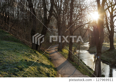 sun shining through trees on path next to river at sunrise. Vysocany district prague 9 Park Zahradky 127969911