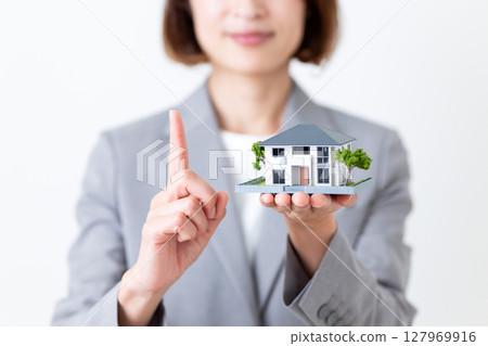 A middle-aged woman in a gray suit holding a model of a house and pointing her index finger up 127969916