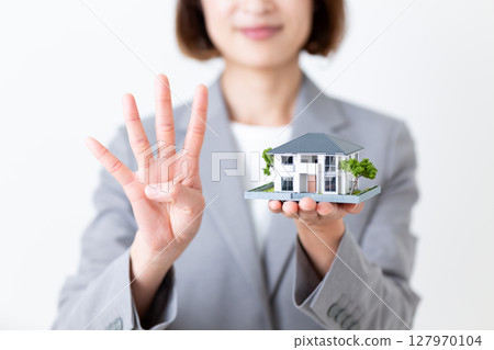 一位身穿灰色西裝的中年婦女，手裡拿著一棟房屋的模型，四根手指向上指著 127970104