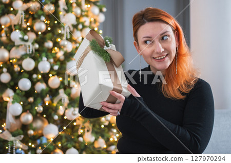 Merry Christmas. Woman unpacking open up gift box near Christmas tree. Girl in living room with Christmas tree holding opening gift box with surprise face. Woman celebrating Christmas eve at home Merry Christmas. Woman unpacking open up gift box near Christmas tree. Girl in living room with Christmas tree holding opening gift box with surprise face. Woman celebrating Christmas eve at home 127970294
