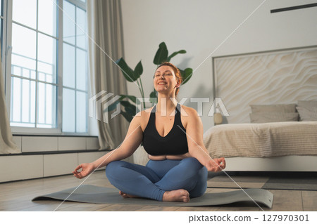 Yoga mindfulness meditation. Woman practicing yoga in living room at home. Plus size female sitting in lotus pose meditating smiling relaxing indoor. Girl doing breathing practice. Yoga at home 127970301
