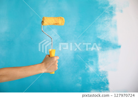close up of female hand holding paint yellow roller over blue background-repair, construction and building tools concept. Convenient and versatile tool for painting walls. close up of female hand holding paint yellow roller over blue background-repair, construction and building tools concept. Convenient and versatile tool for painting walls. 127970724