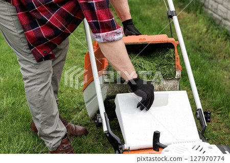 Gardener man in casual clothes carries plastic lawn mower box with cut grass of modern gasoline lawn mower at backyard of house at spring day. Using grass clippings for compost or vegetable garden 127970774
