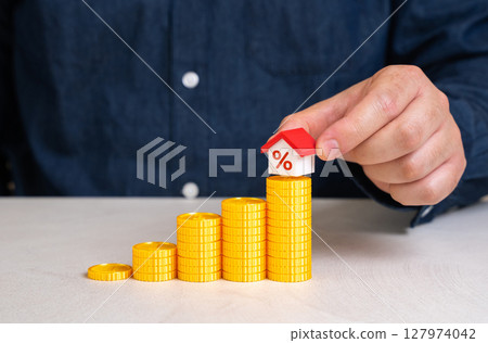 A man places a house on top of a staircase made of coins. Saving for a house. Down payment and opening a mortgage. Financial planning, real estate investment, and overcoming barriers to homeownership A man places a house on top of a staircase made of coins. Saving for a house. Down payment and opening a mortgage. Financial planning, real estate investment, and overcoming barriers to homeownership 127974042