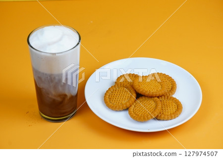 A refreshing glass of iced coffee with a foamy top and a plate of golden-brown biscuits, all against a vibrant orange backdrop. 127974507