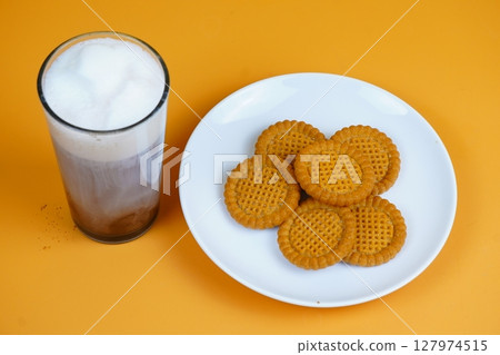 A refreshing glass of iced coffee with a foamy top and a plate of golden-brown biscuits, all against a vibrant orange backdrop. A refreshing glass of iced coffee with a foamy top and a plate of golden-brown biscuits, all against a vibrant orange backdrop. 127974515