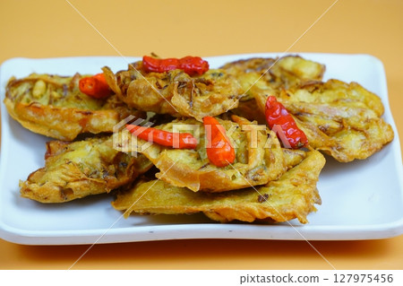 A plate of Indonesian fried vegetable snacks or bakwan topped with red chilies, set against an orange background. 127975456