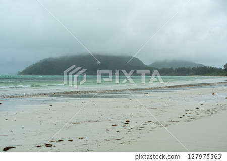 Tropical beach on Praslin island in Seychelles with misty hills, cloudy sky, wet sand, turquoise ocean 127975563
