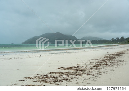 Tropical beach on Praslin island in Seychelles with misty hills, cloudy sky, wet sand, turquoise ocean 127975564