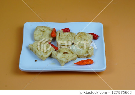 A close up of several pieces of fried tofu, a common Indonesian snack, served on a white plate with red chilies, against an orange background. 127975663