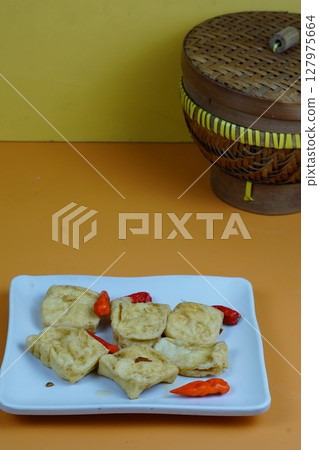 A close up of several pieces of fried tofu, a common Indonesian snack, served on a white plate with red chilies, against an orange background. 127975664