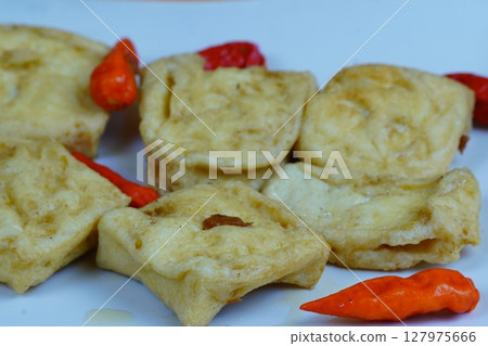 A close up of several pieces of fried tofu, a common Indonesian snack, served on a white plate with red chilies, against an orange background. 127975666