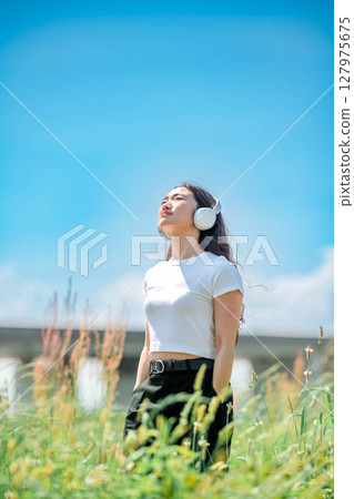 一位身穿白色 T 恤的年輕女子在藍天下戴著耳機聽音樂 127975675