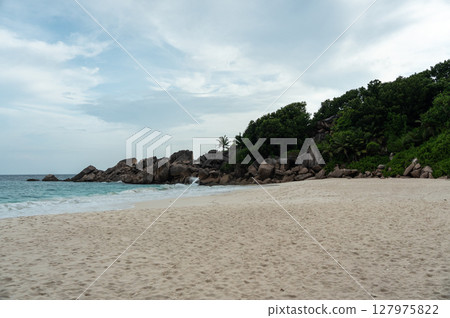 Granite boulders and dense tropical vegetation line the beach on La Digue island Granite boulders and dense tropical vegetation line the beach on La Digue island 127975822